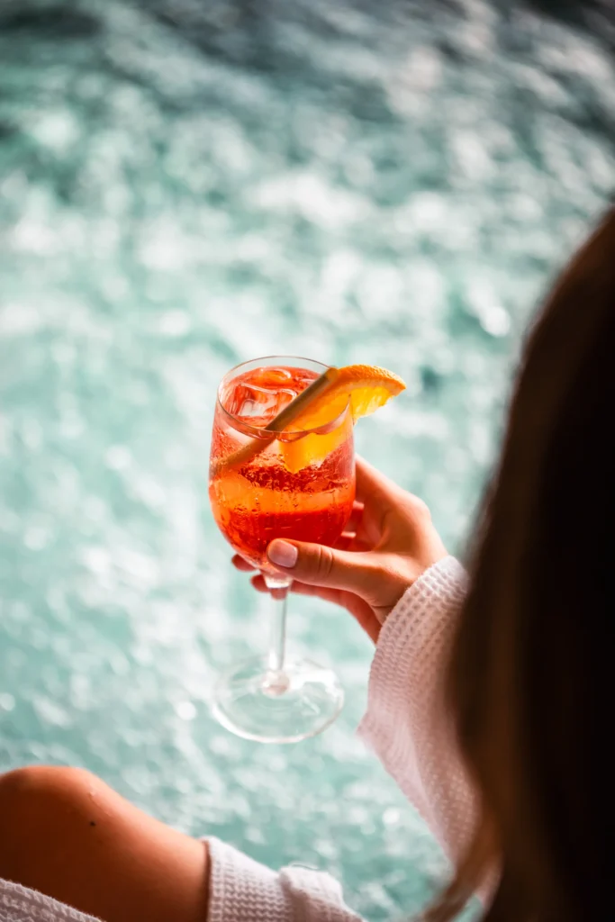 En person i vit morgonrock håller ett glas apelsincocktail med is, sugrör och en apelsinskiva som garnityr, med en suddig bakgrund av blått vatten.