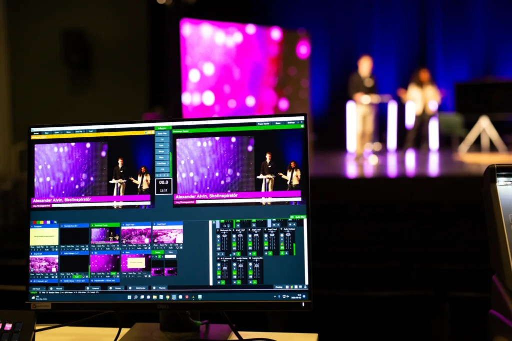 En datorskärm visar en direktsänd videoflöde och kontroller för en scenhändelse, med två suddiga presentatörer som står vid podier i bakgrunden under lila belysning.