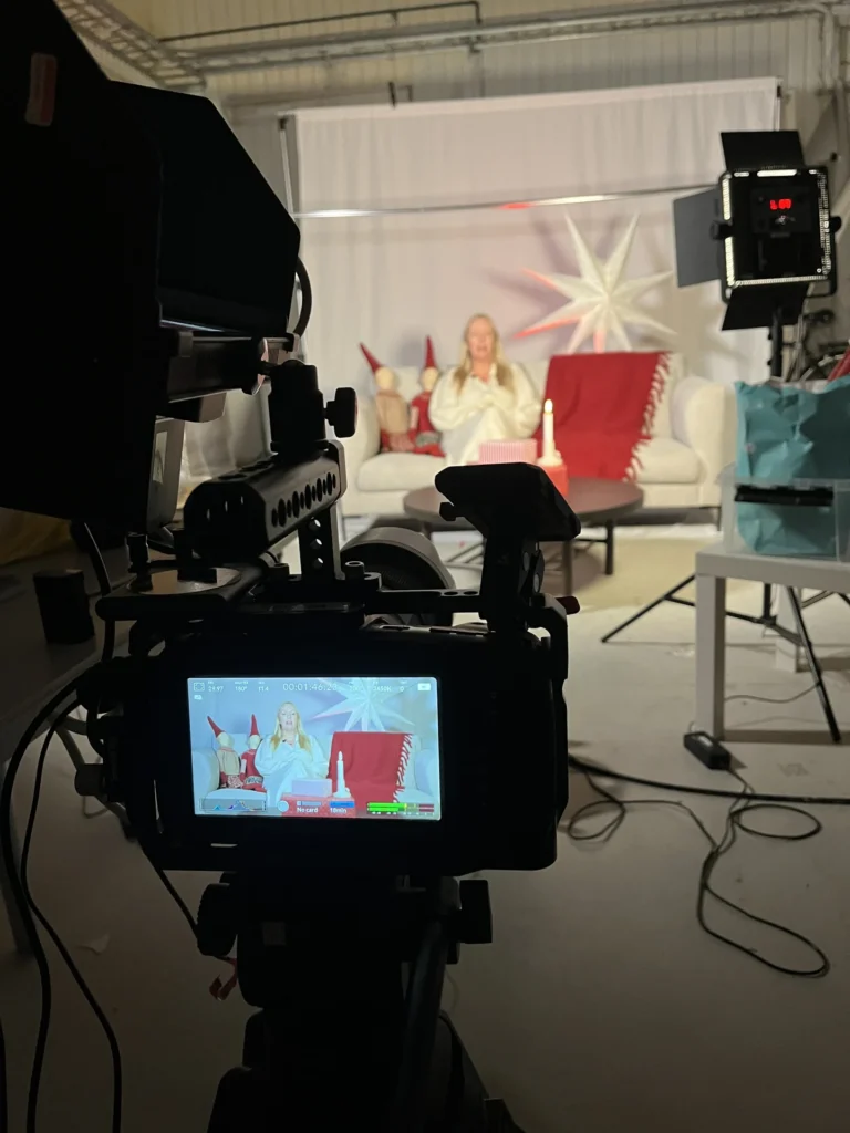 En kamera filmar en festklädd person som sitter i en vit soffa med röda kuddar och stora vita stjärndekorationer, i en starkt upplyst studio - perfekt för en inspirerande filmproduktion för företag.