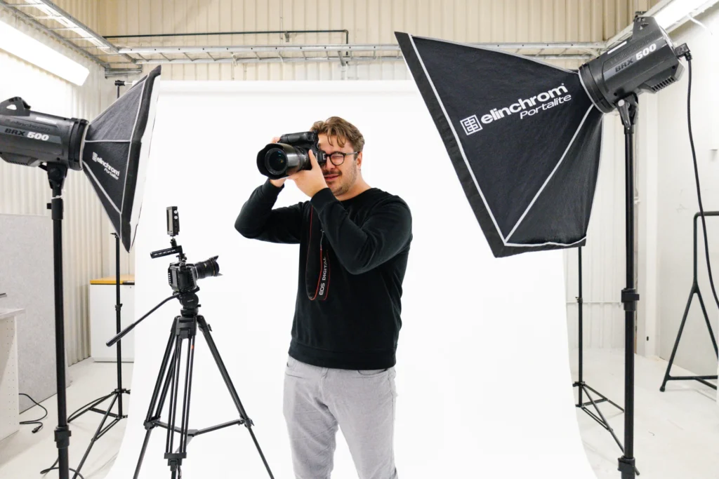 En man med glasögon står i en fotostudio och håller en kamera mot sitt öga. Han är omgiven av professionell belysningsutrustning och en vit bakgrund, med en annan kamera monterad på ett stativ i närheten.