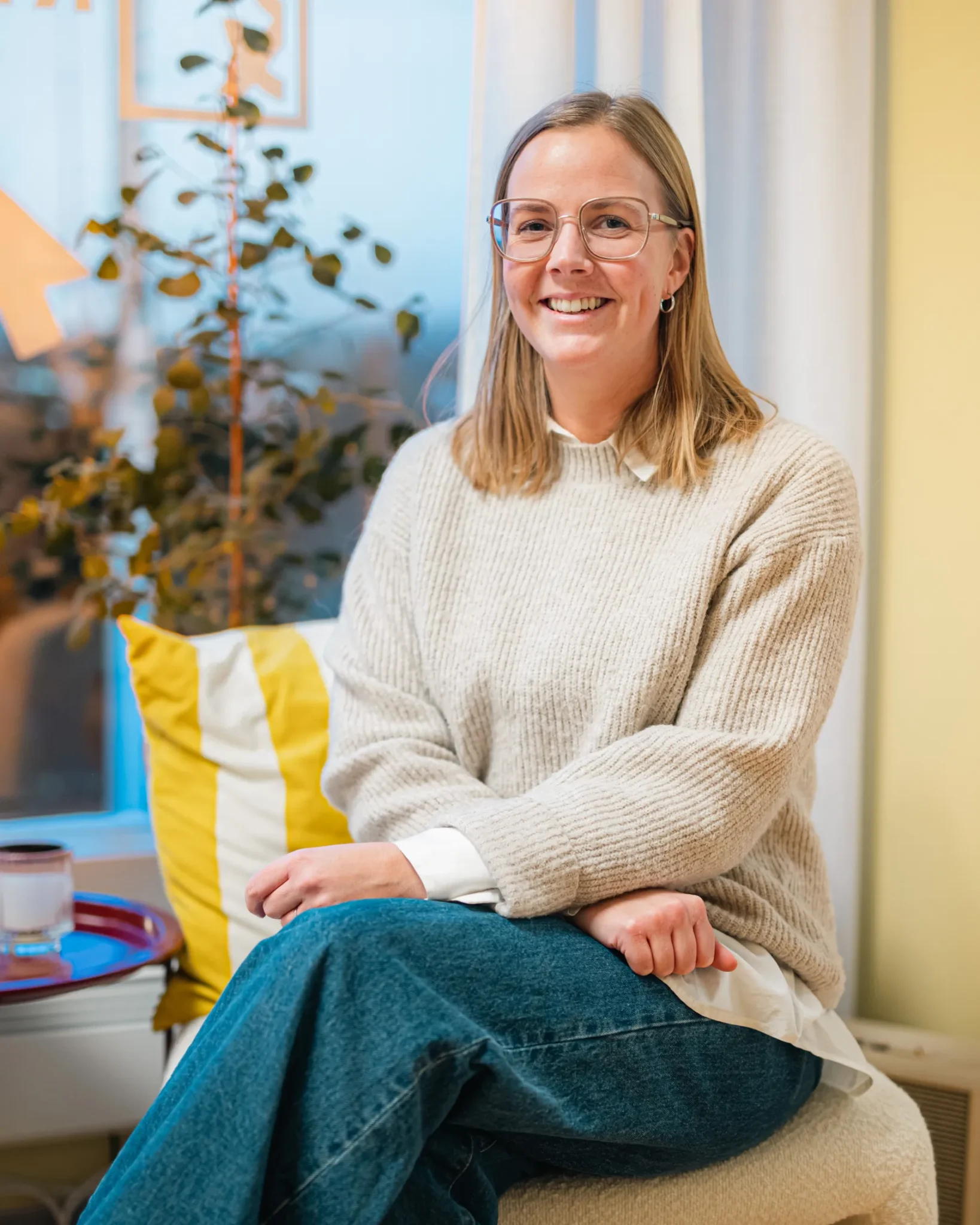 En kvinna med axellångt blont hår och glasögon, klädd i en beige tröja och blå jeans, sitter leende på en stol i ett mysigt rum med en randig kudde och en växt vid fönstret.