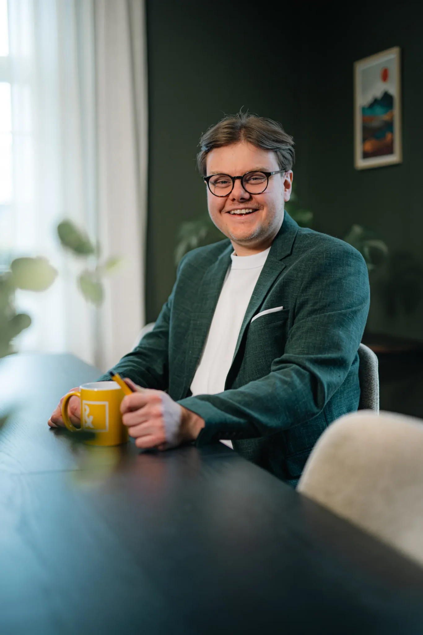 En leende man med glasögon och grön kavaj sitter vid ett bord och håller i en gul mugg. I den svagt upplysta bakgrunden finns växter och en inramad tavla.