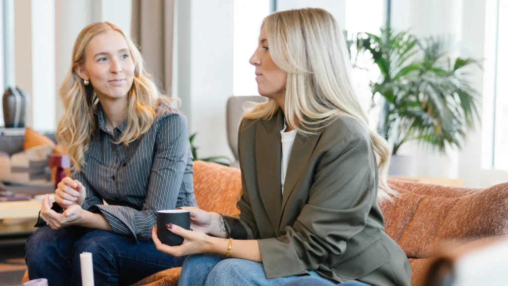 Två kvinnor med blont hår sitter i en orange soffa på Reklam & Co byrå i Motala med koppar i handen och småpratar. De verkar avslappnade och engagerade, med växter och stora fönster i bakgrunden som antyder en modern inomhusmiljö.