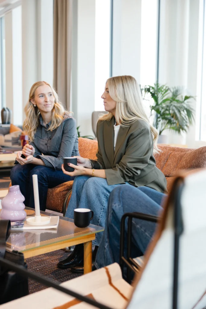 I ett modernt, ljust rum på Reklam & Co byrå i Motala sitter två kvinnor i en orange soffa och samtalar. Den ena håller i en kopp och bär en grön kavaj, den andra har en randig skjorta och jeans. Framför dem står ett soffbord.