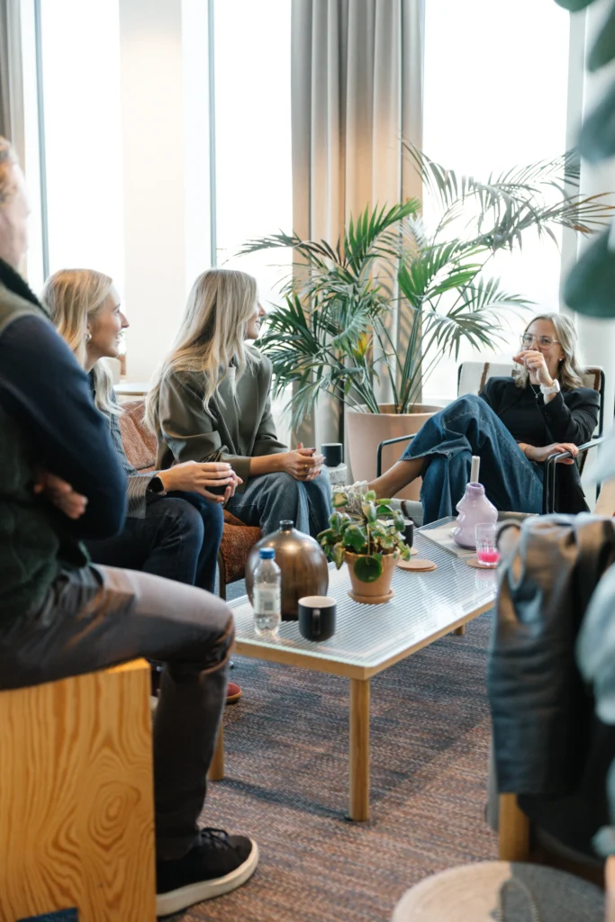 Fyra personer sitter och småpratar runt ett soffbord i Reklam & Co byrå i Motalas ljusa, moderna kontor med stora fönster och växter. De ler och håller i koppar, vilket tyder på en vänlig och avslappnad konversation.