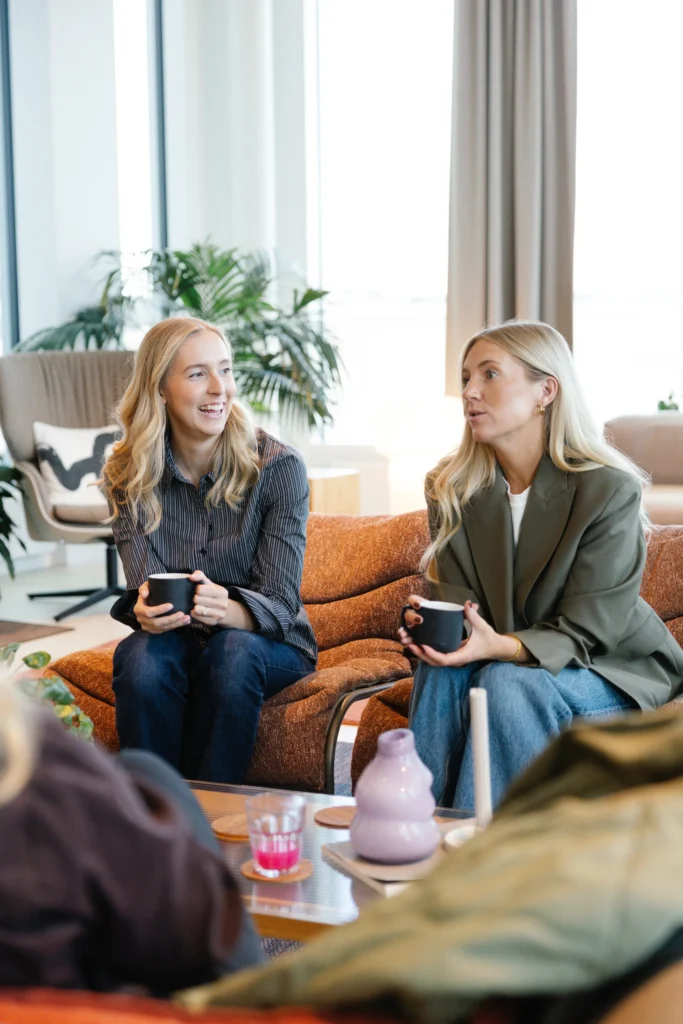 Två kvinnor med långt blont hår sitter i en orange soffa med svarta muggar i handen och småpratar. Den ena ler medan den andra ser fundersam ut. I den mysiga Motalamiljön finns växter och ett bord med koppar, perfekt för kreativa samtal på Reklam & Co byrå i Motala.