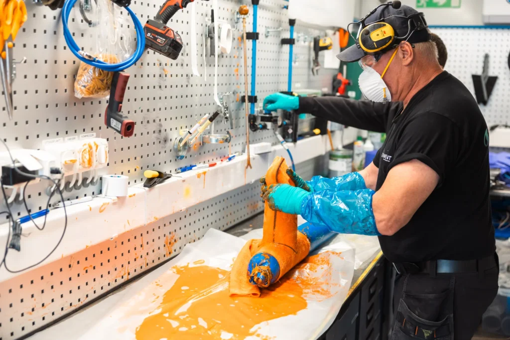 En person iförd handskar, mask och hörselskydd arbetar vid en bänk och hanterar ett orangefärgat föremål med verktyg och utrustning på väggen i en verkstad. Orange vätska stänker ut på arbetsytan.