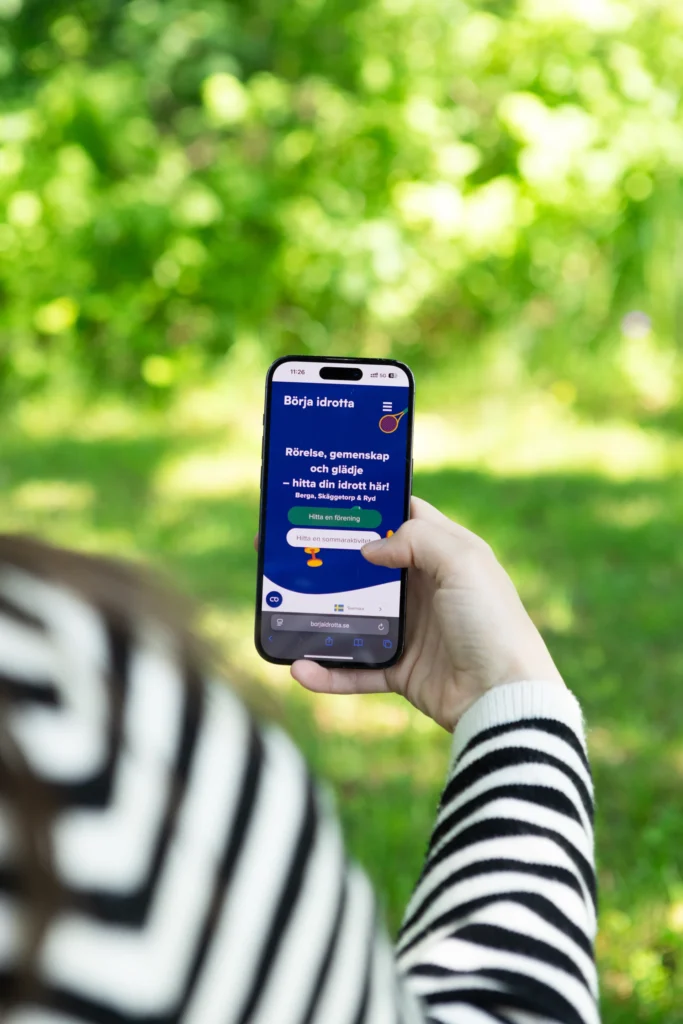 En person i svartvitrandig tröja håller i en smartphone utomhus och visar en svensk webbplats om att börja med sportaktiviteter. Bakgrunden är grön och suddig med lövverk.