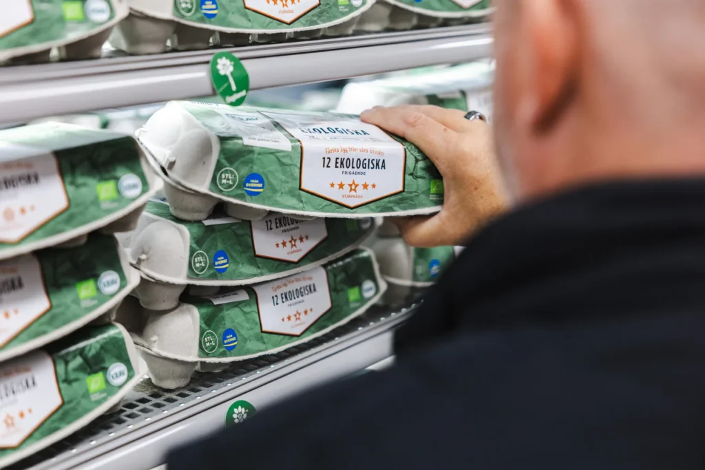 En person väljer en kartong med ekologiska ägg märkta 12 Ekologiska från en hylla i en livsmedelsbutik med liknande äggkartonger.