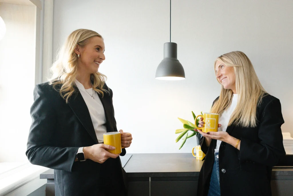 Två kvinnor med långt blont hår, klädda i svarta kavajer och vita toppar, står i ett modernt kök med gula muggar i handen, ler och pratar med varandra. En svart pendellampa hänger ovanför köksbänken.
