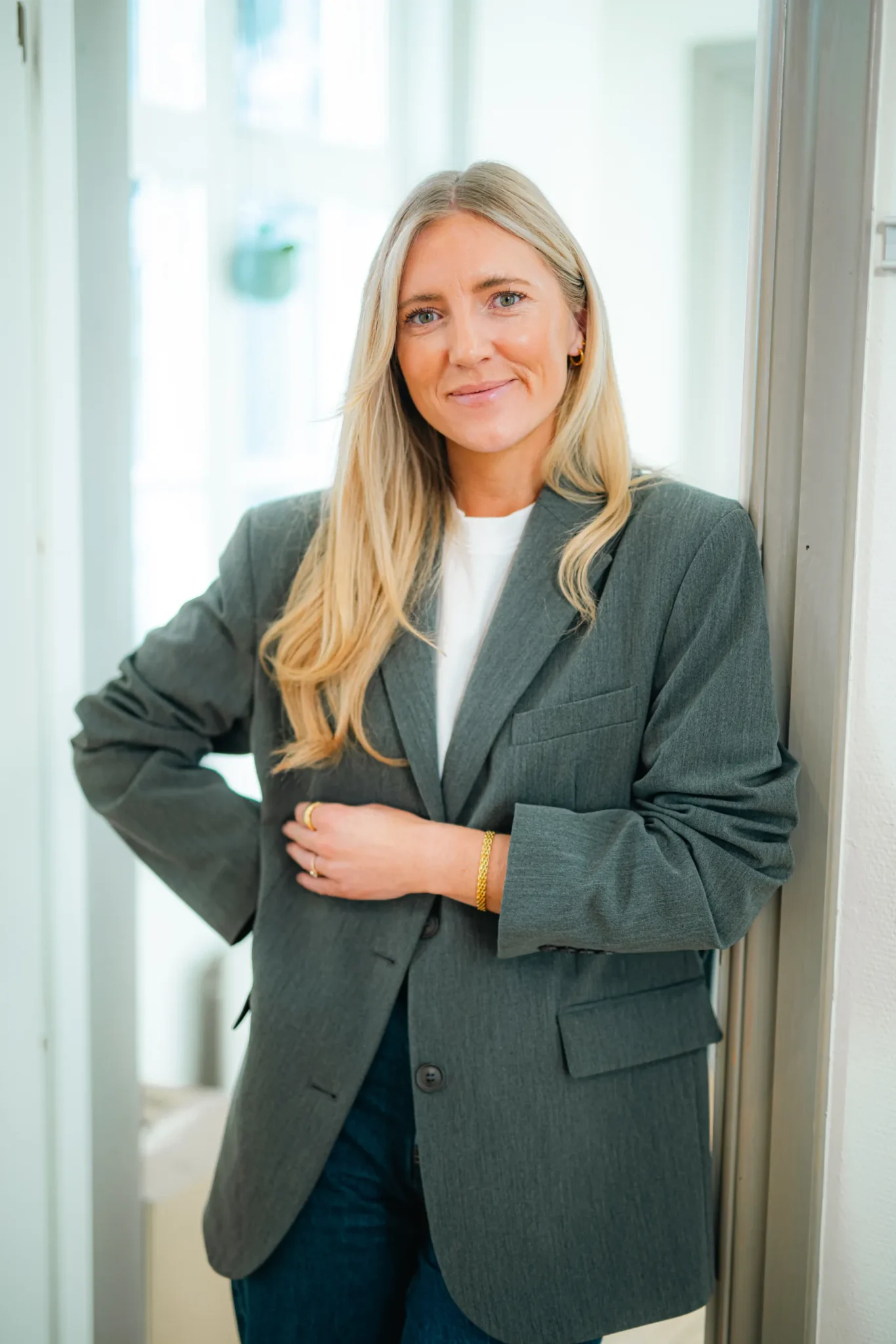 En kvinna med långt blont hår, klädd i en grå kavaj över en vit topp och blå jeans, står inomhus lutad mot en vägg och ler mjukt mot kameran.