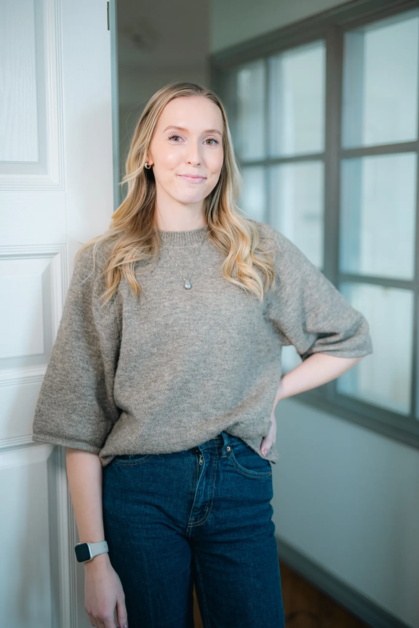 En kvinna med långt blont hår, grå tröja och blå jeans står inomhus och lutar sig avslappnat mot en vit dörr med ena handen på höften och ler mjukt.