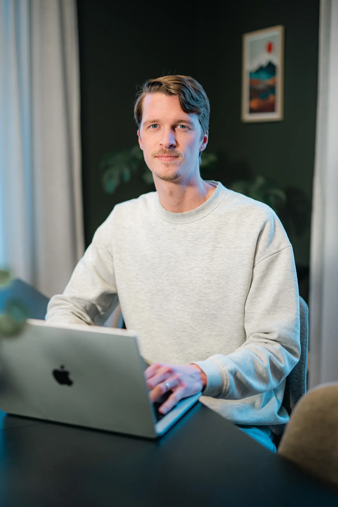 En man med ljusbrunt hår och en ljusgrå sweatshirt sitter vid ett bord med en silverfärgad MacBook laptop och tittar in i kameran. Bakgrunden består av gröna väggar, en växt, gardiner och en inramad tavla.