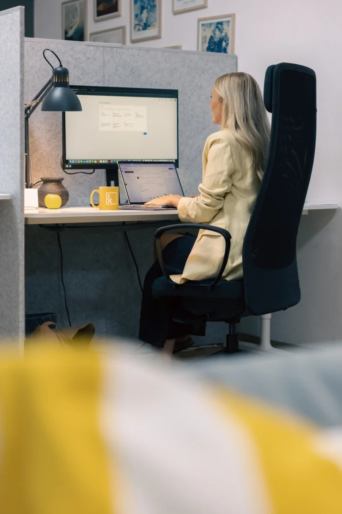 En kvinna med långt blont hår sitter vid ett skrivbord i ett kontorslandskap och arbetar med betald annonsering för företag på sin bärbara dator och stora bildskärm. I bakgrunden syns en lampa, en gul mugg och kontorsinredning.