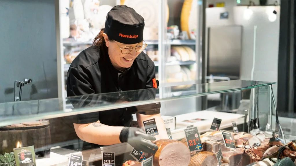 En leende Hemköp-delikatessarbetare i uniform och handskar står bakom en glasdisk och skivar och arrangerar skickligt olika köttsorter för utställning i en ljus, modern butik.