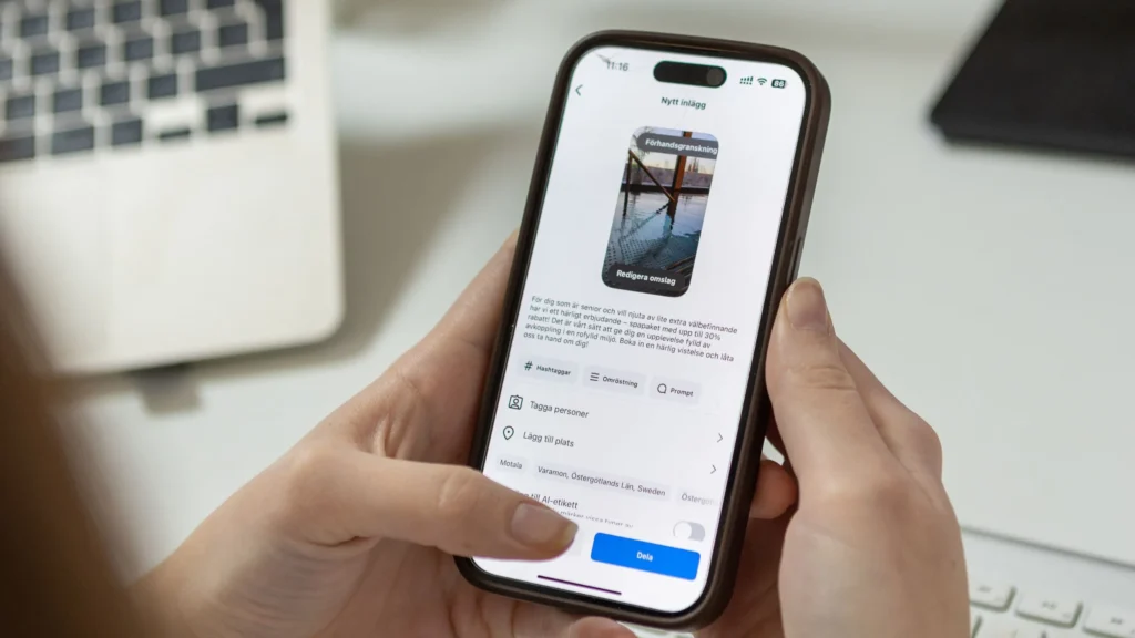 En person håller i en smartphone och redigerar ett inlägg på sociala medier med ett foto av en pir över vatten. En bärbar dator och ett tangentbord syns i den suddiga bakgrunden.