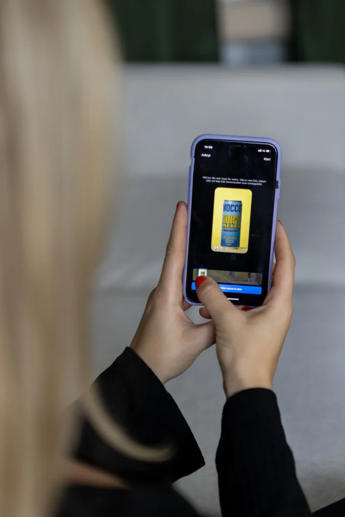 En person med blont hår håller i en smartphone och tittar på en bild av en gul energidrycksburk märkt NOCCO på skärmen. Personens händer och telefon är i fokus; bakgrunden är suddig.