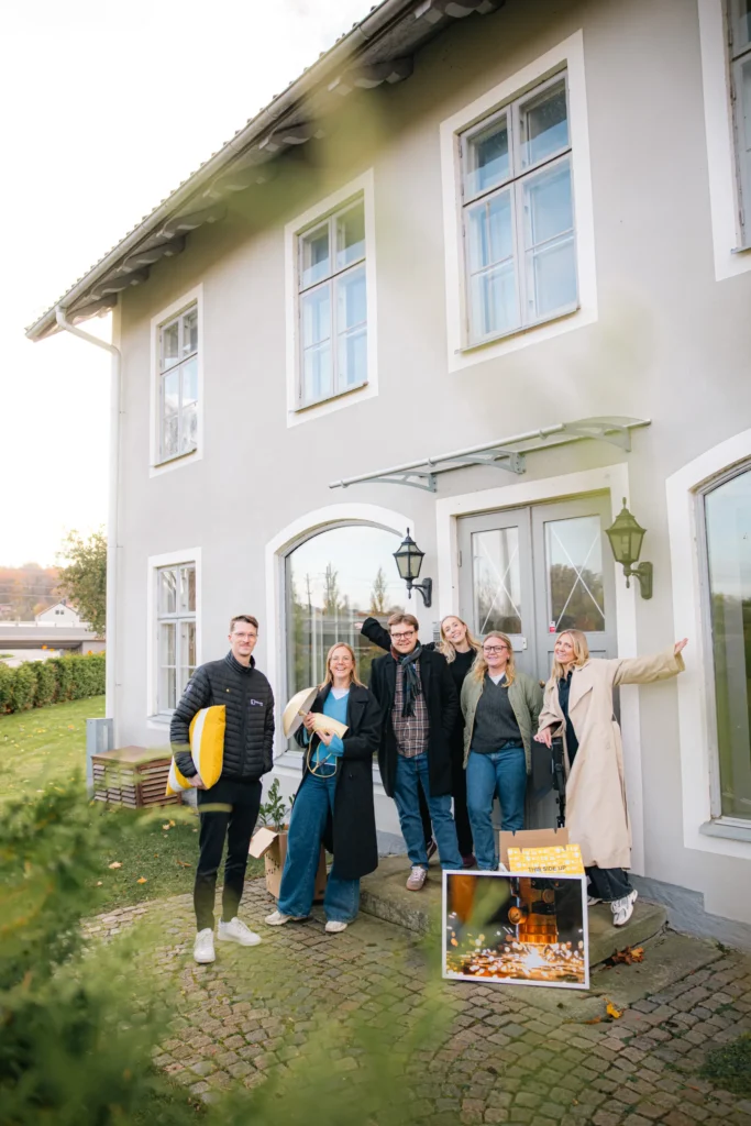 Fem personer står leende framför ett ljusgrått hus med stora fönster. De verkar glada och håller i olika föremål, bland annat en gul kudde och ett inramat foto som ligger på marken nära dörröppningen.
