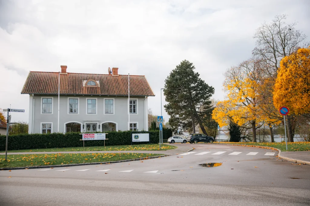 Ett stort, vitt tvåvåningshus med rött tegeltak står i ett gathörn, omgivet av gröna häckar och höstträd med gula löv. Bilar är parkerade i närheten och en vattenpöl ligger på vägen.