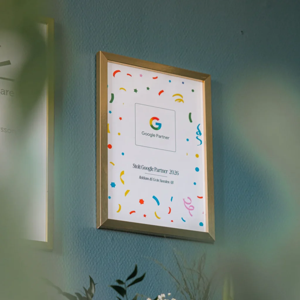 Ett inramat Google Partner-certifieringspris med färgglada konfettimotiv hänger på en blå vägg ovanför en vas med blommor, bland annat blåsippor och en rosa blomma. Vissa bladverk är suddiga i förgrunden.