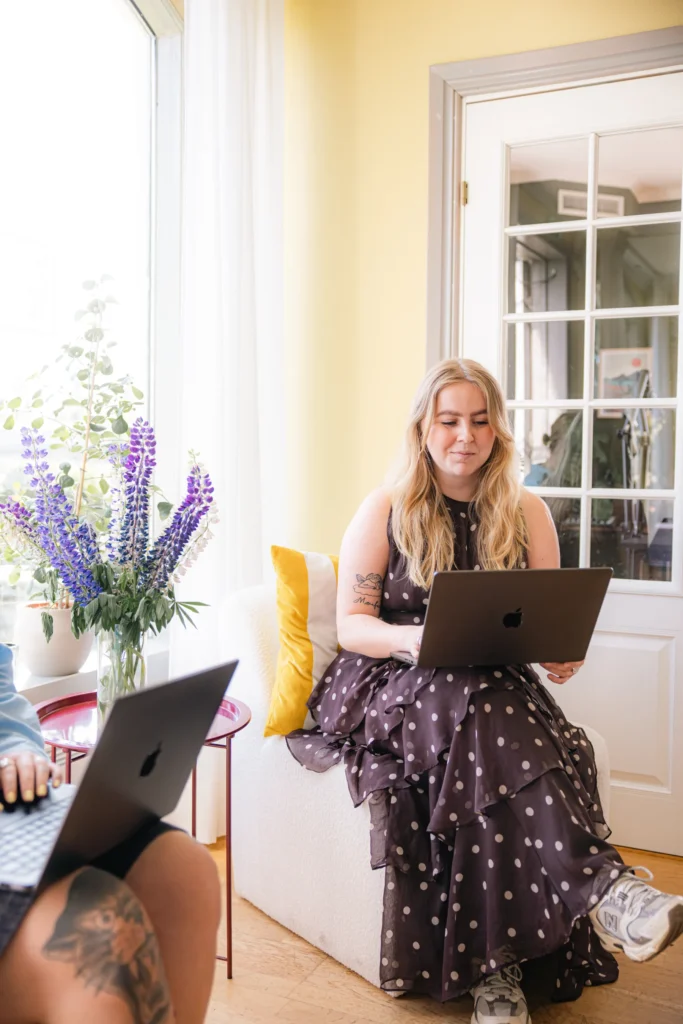 En kvinna i en prickig klänning sitter på en vit stol och arbetar på sin bärbara dator, kanske för att förbereda sig inför sin Google Partner-certifiering, bredvid en vas med lila blommor i ett ljust rum med gula väggar och ett stort fönster.