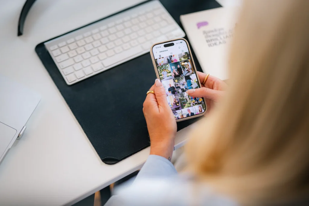 En person håller en smartphone i handen och bläddrar igenom ett fotogalleri. På skrivbordet syns ett datortangentbord, en musmatta och en del av en bärbar dator. Miljön är ljus och välordnad.