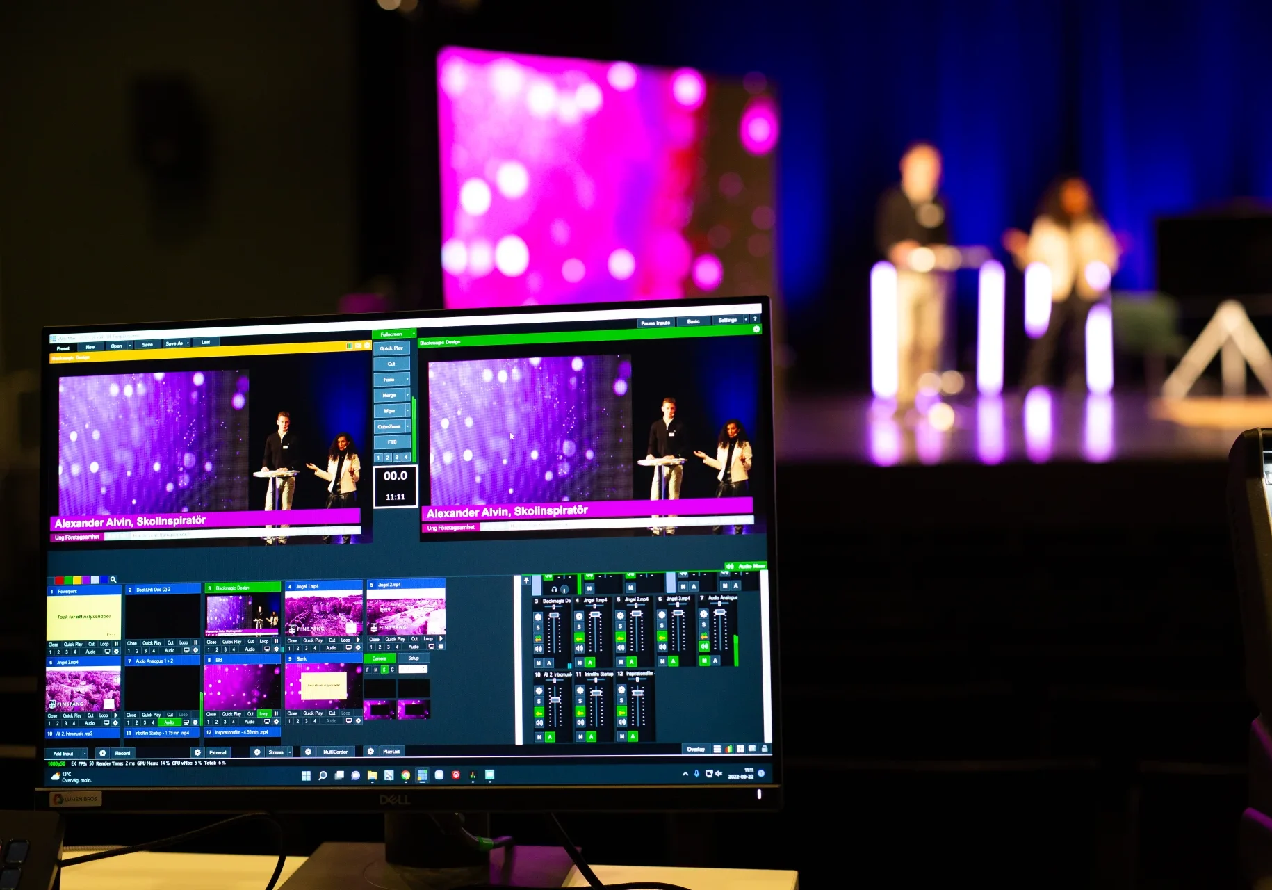 Livesändning - Reklam & Co En datorskärm visar en direktsänd videoflöde och kontroller för en scenhändelse, med två suddiga presentatörer som står vid podier i bakgrunden under lila belysning.