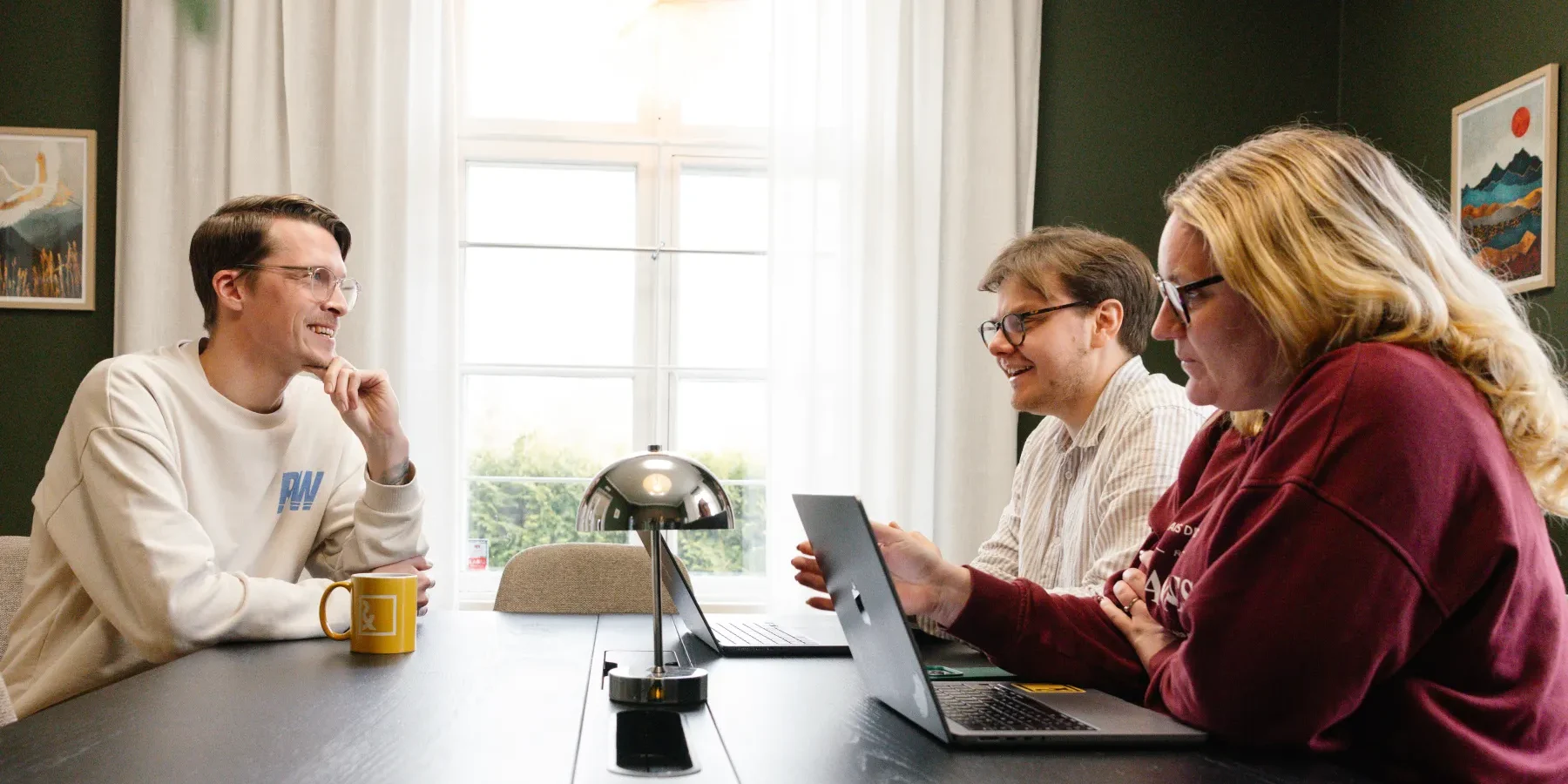 Tre personer sitter runt ett bord i ett väl upplyst rum med bärbara datorer, ler och diskuterar AI och SEO 2026. I bakgrunden syns ett fönster med vita gardiner och inramad konst.