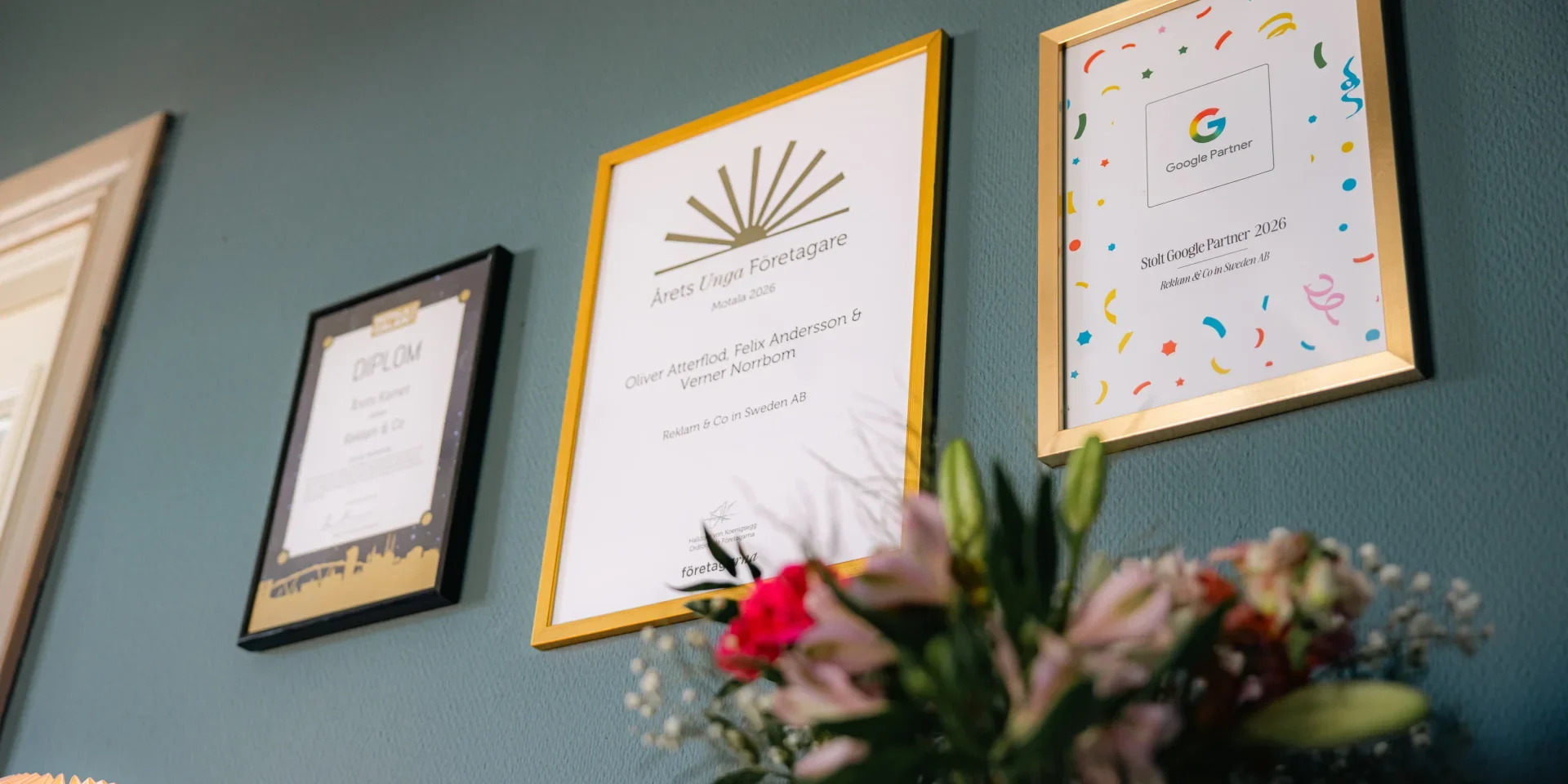 Tre inramade certifikat hänger på en grön vägg ovanför en blombukett, varav ett med ett Google Partner-certifieringsmärke och ett annat med en utmärkelse för ett företag eller en verksamhet.