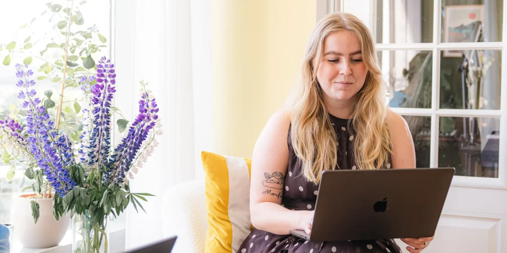En kvinna i en prickig klänning sitter på en vit stol och arbetar på sin bärbara dator, kanske för att förbereda sig inför sin Google Partner-certifiering, bredvid en vas med lila blommor i ett ljust rum med gula väggar och ett stort fönster.