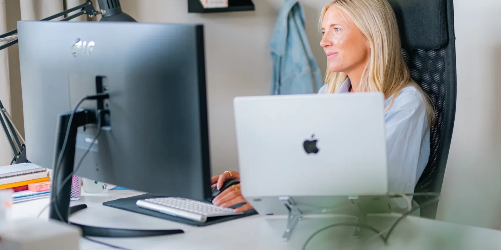 AI-transparenslagen En kvinna med långt blont hår sitter vid ett skrivbord i ett modernt kontor och arbetar vid en stationär dator och en bärbar dator. Hon är helt upptagen av sina skärmar. Arbetsplatsen är ljus och välorganiserad, med ett trådgaller på väggen bakom henne.
