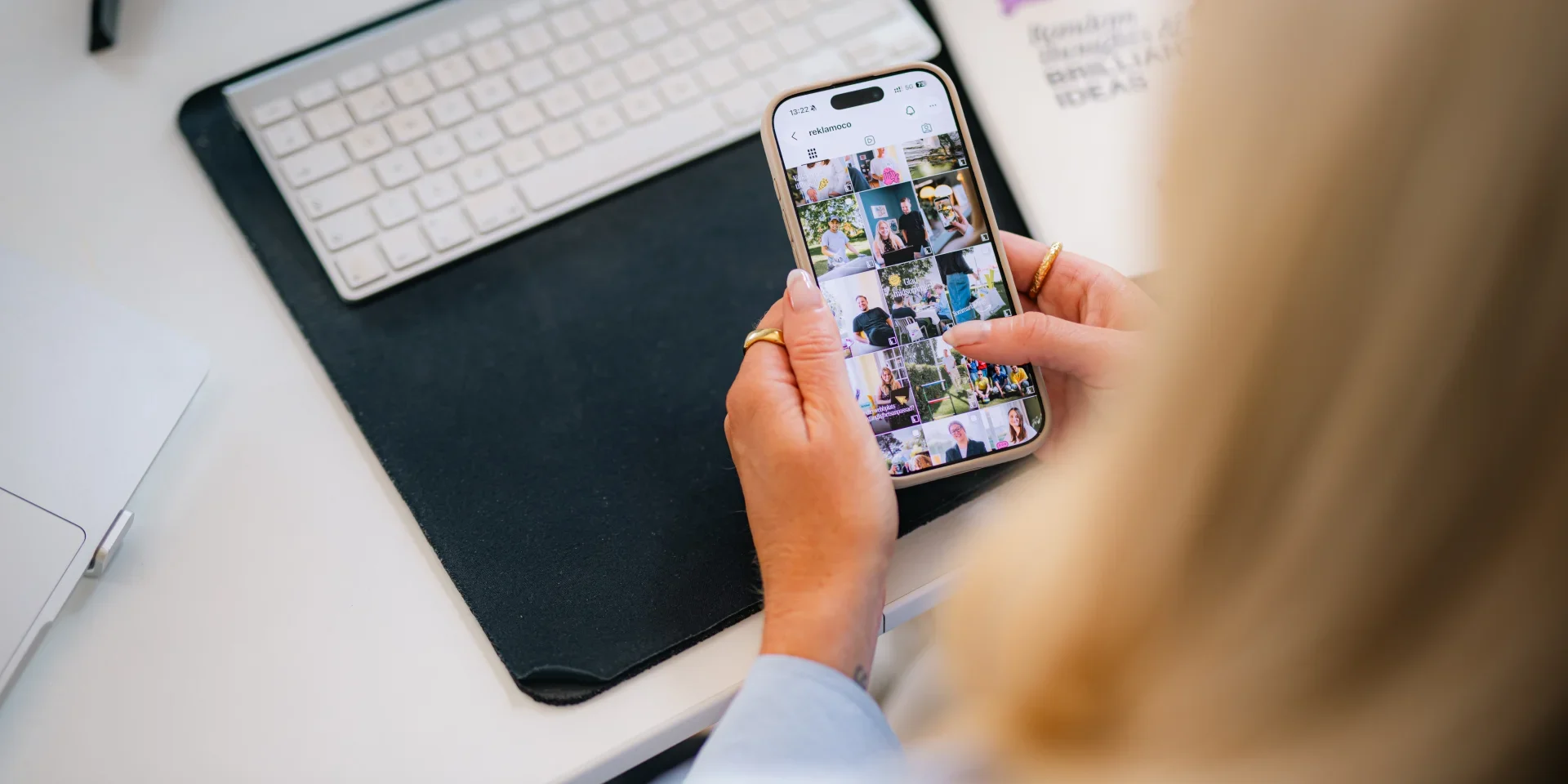 AI på sociala medier En person håller en smartphone i handen och bläddrar igenom ett fotogalleri. På skrivbordet syns ett datortangentbord, en musmatta och en del av en bärbar dator. Miljön är ljus och välordnad.