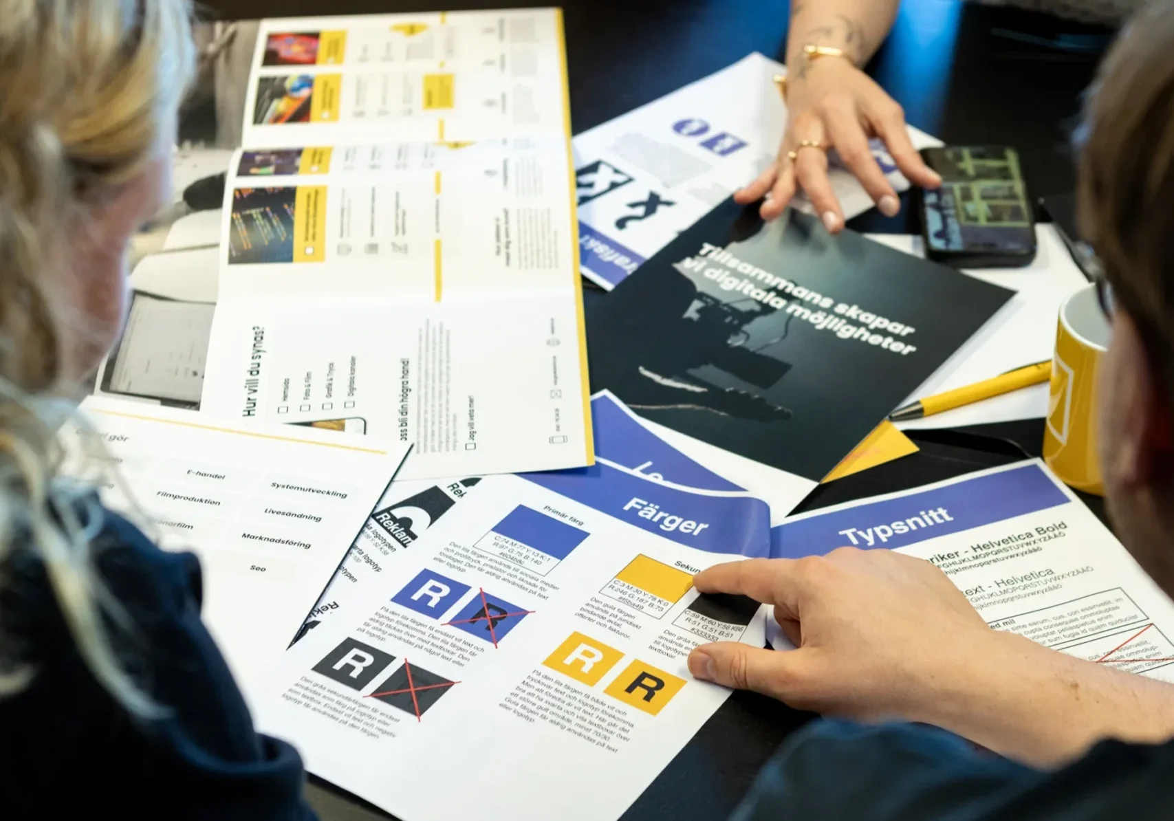 Layout & logotyper - Rekalm & Co Fyra personer sitter runt ett bord och diskuterar tryckta dokument och broschyrer med diagram, text och färgprover, vilket tyder på ett möte eller en gemensam arbetssession inför en livesändning. Endast deras armar och händer är synliga.