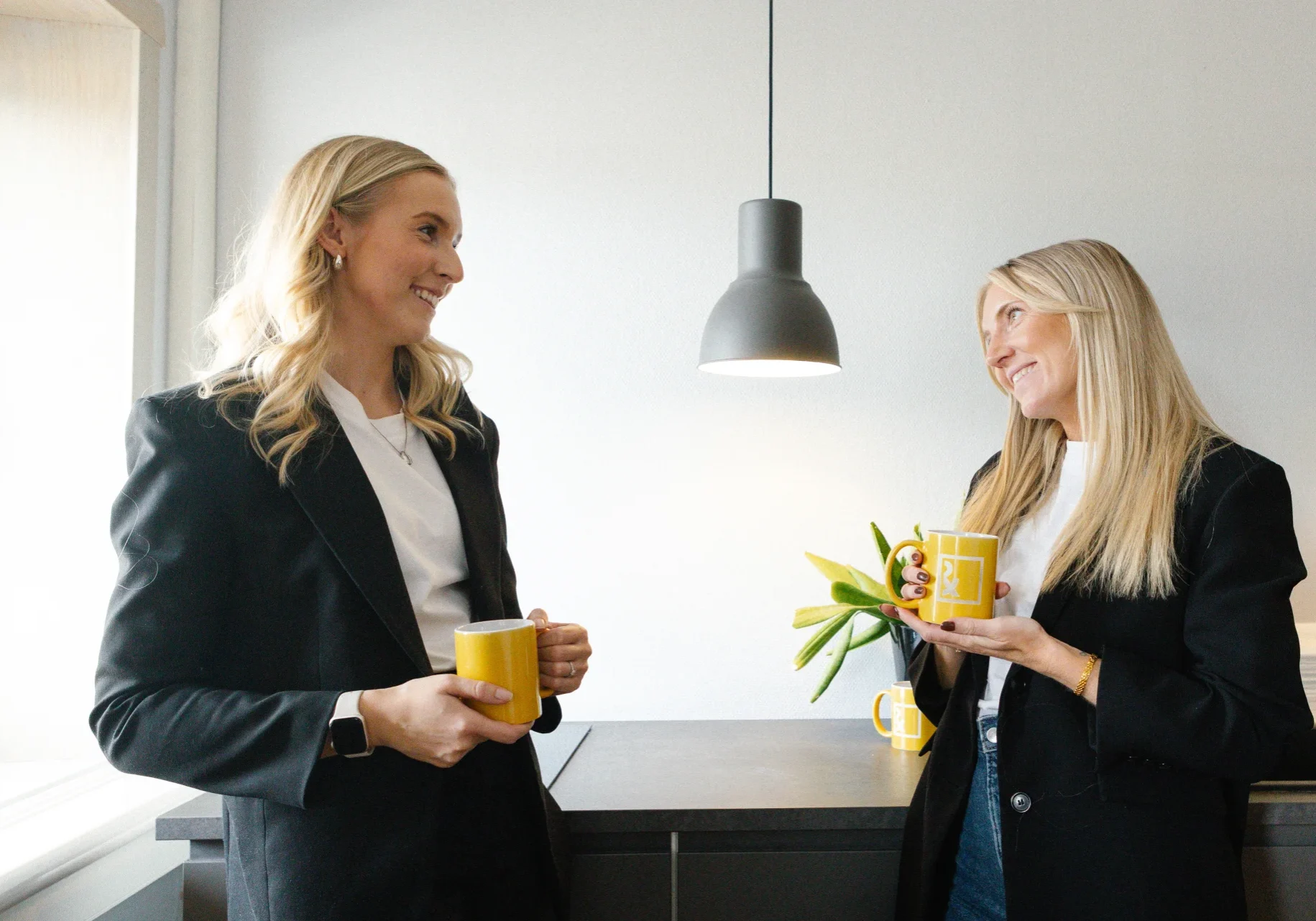 Sofia & Sandra - Reklam & Co Två kvinnor med långt blont hår, klädda i svarta kavajer och vita toppar, står i ett modernt kök med gula muggar i handen, ler och pratar med varandra. En svart pendellampa hänger ovanför köksbänken.