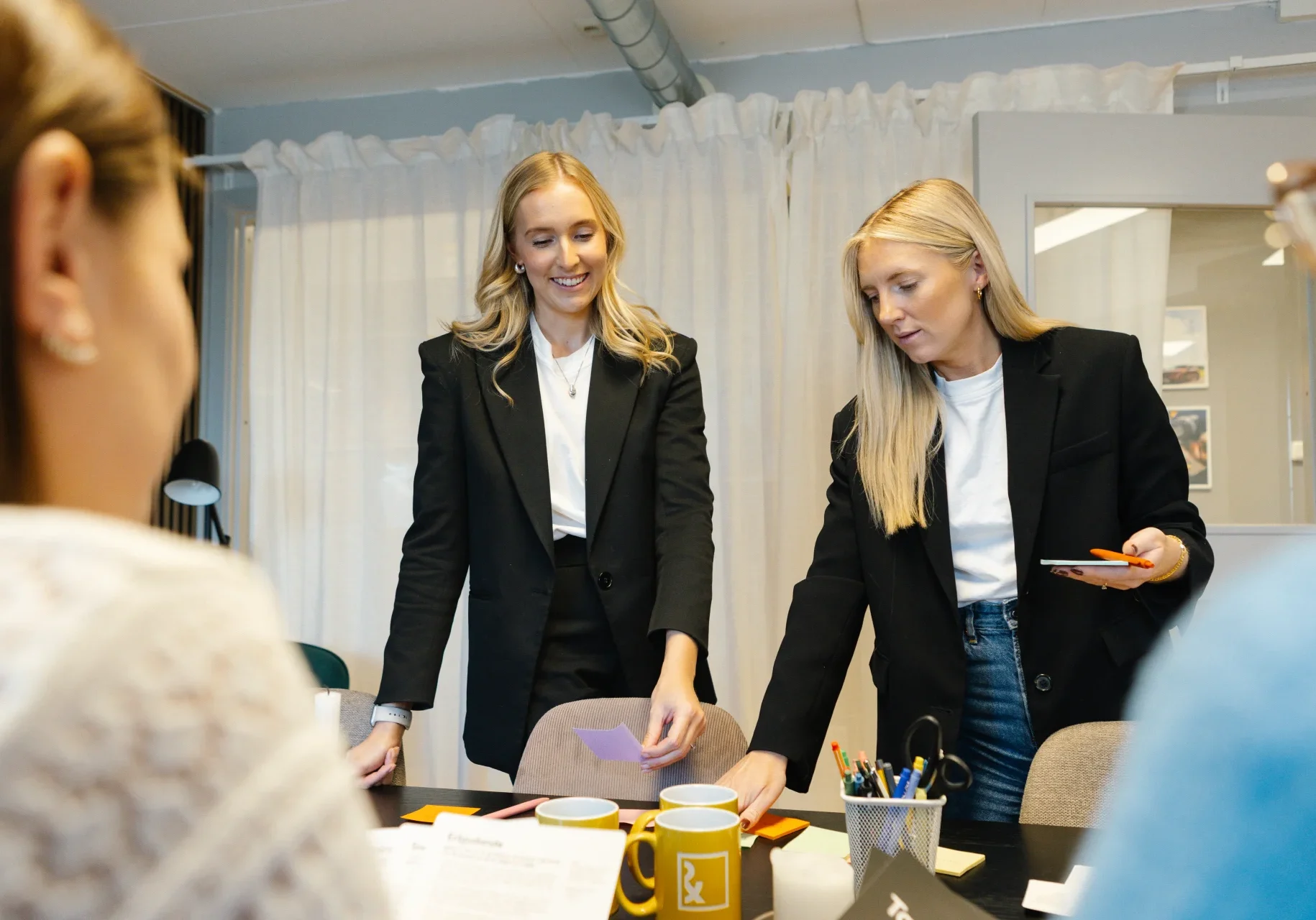 Din marknadskoordinator - Reklam & Co Två kvinnor i kavaj står vid ett bord med papper, muggar och kontorsmaterial och samarbetar i ett ljust, modernt kontor medan två andra personer sitter i närheten.