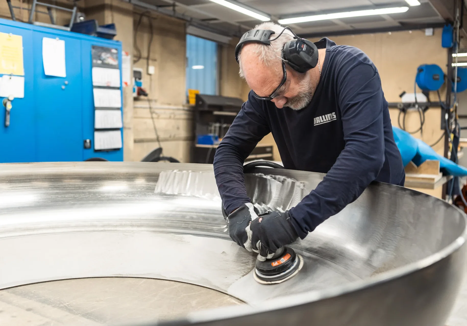 En man med skyddsglasögon, handskar och hörselkåpor använder en slipmaskin för att polera kanten på en stor metallring i en industriell verkstad. Blå skåp och verktyg syns i bakgrunden.