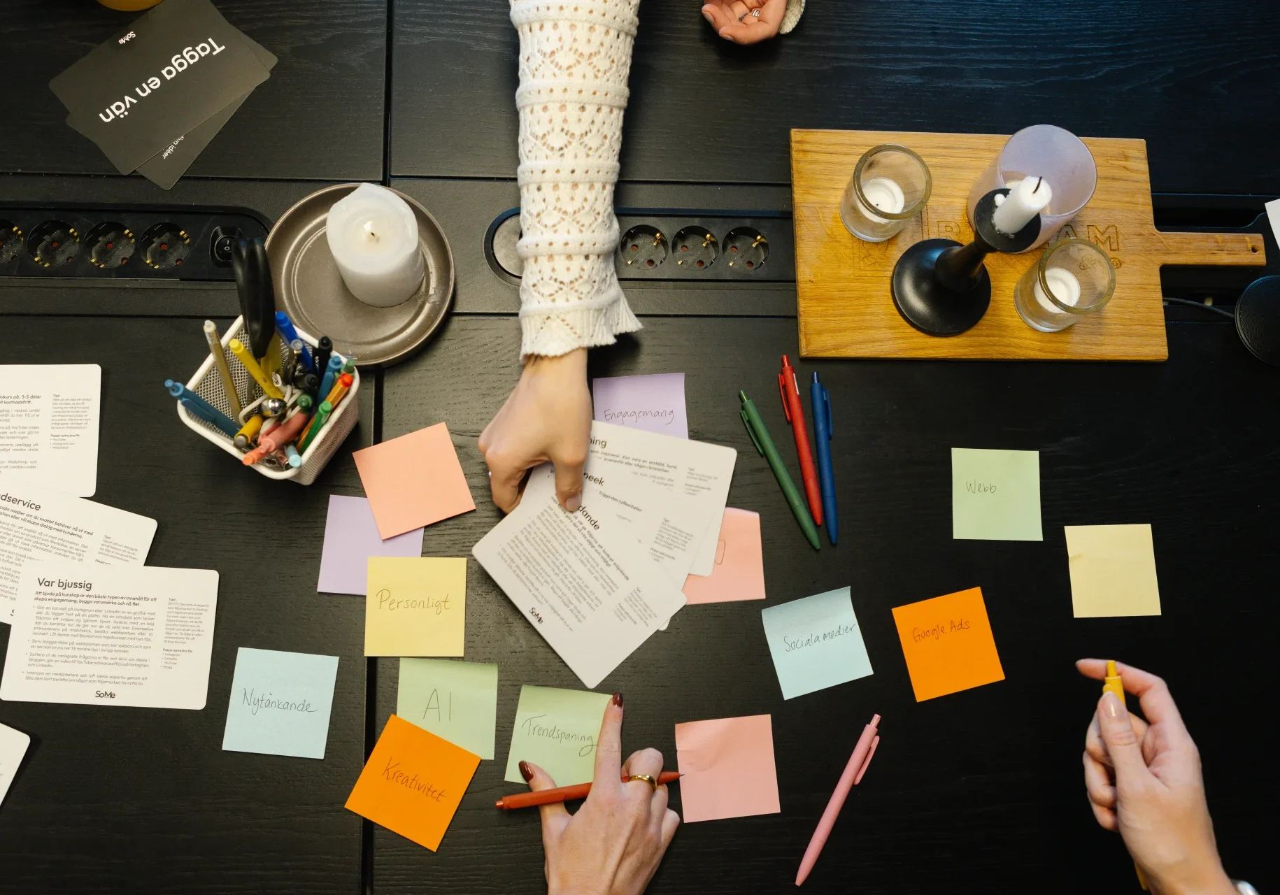 Analys Tre personer arbetar vid ett bord som är täckt med post-it-lappar, papper, pennor och ljus. En person pekar på ett dokument, medan de andra skriver på lappar och håller i pennor, vilket tyder på en gemensam planeringssession.