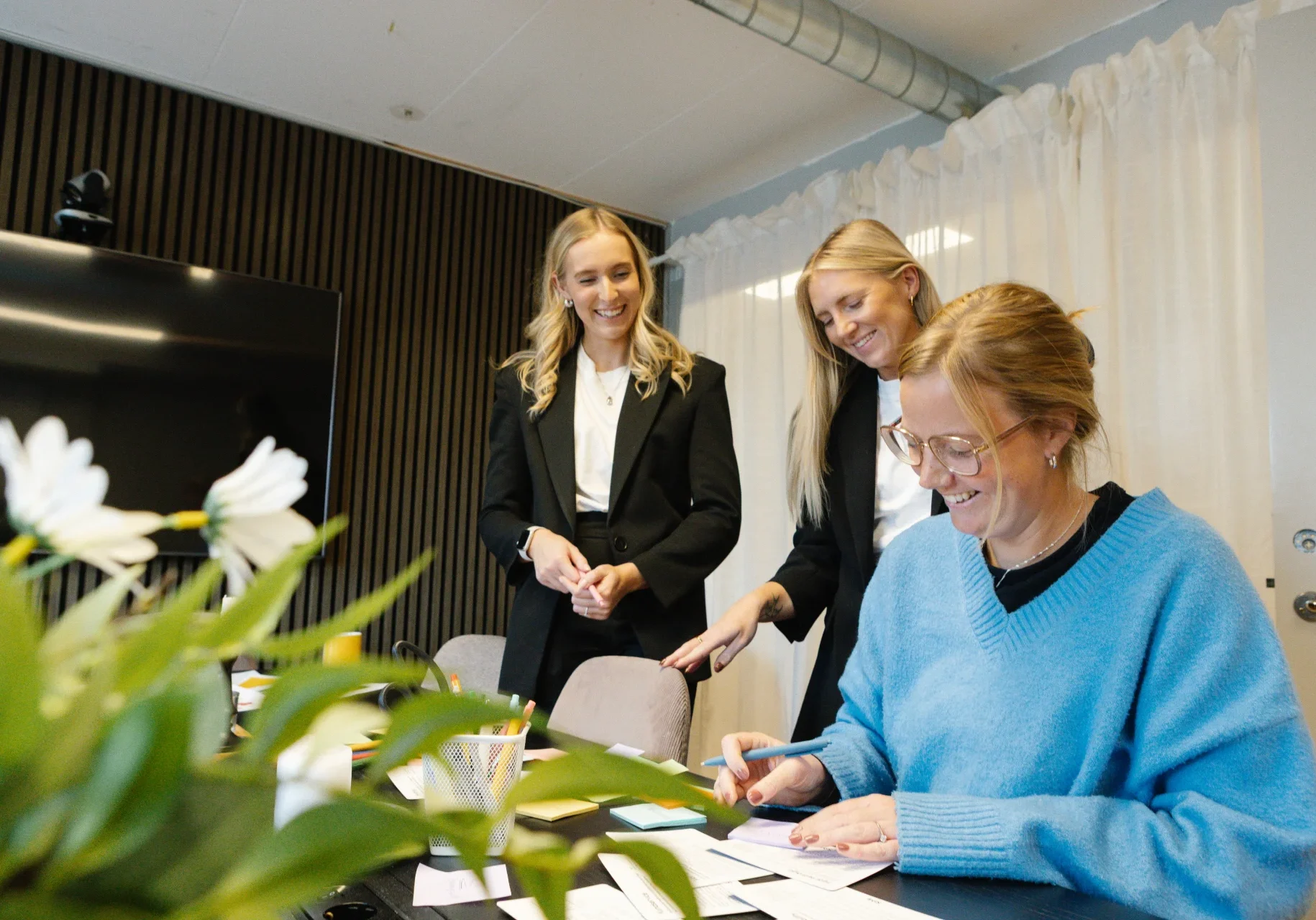 Betald annonsering - Rekalm & Co Tre kvinnor befinner sig i ett mötesrum. En kvinna i blå tröja sitter och skriver på ett papper, medan två andra kvinnor i svarta kavajer står bakom henne och ler. I förgrunden syns en växt och i bakgrunden vita gardiner.