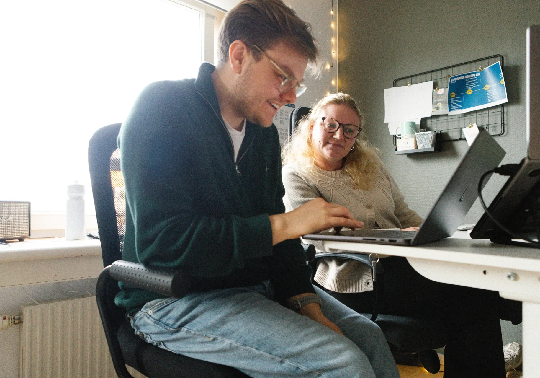 Hemsideproduktion - Reklam & Co En man och en kvinna sitter vid ett skrivbord på ett kontor och tittar båda på en laptopskärm. Mannen skriver medan kvinnan tittar uppmärksamt på. I bakgrunden syns ett fönster och en väggorganisatör.