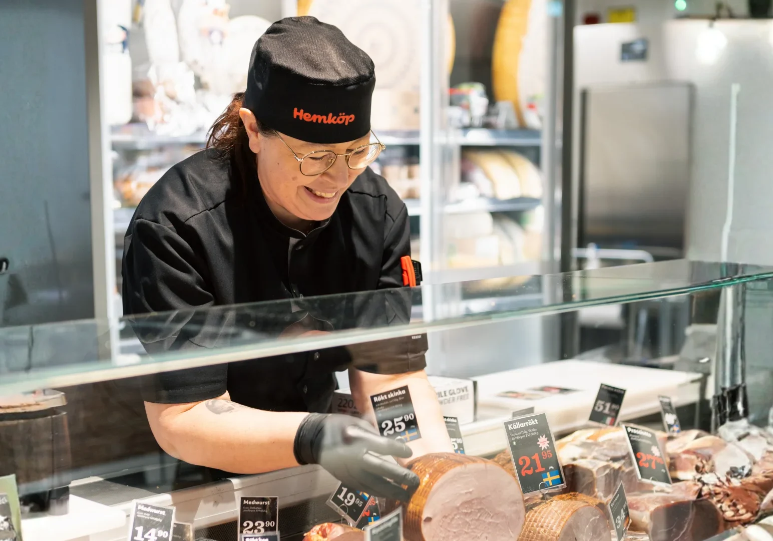 En leende Hemköp-delikatessarbetare i uniform och handskar står bakom en glasdisk och skivar och arrangerar skickligt olika köttsorter för utställning i en ljus, modern butik.